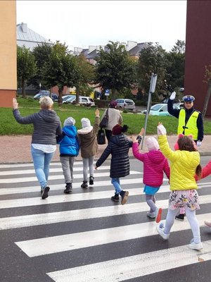 dzieci wraz z wychowawczynią przechodzą przez przejście dla pieszych, z boku stoi umundurowany policjant