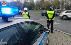 radiowóz oznakowany z włączonymi sygnałami świetlnymi oraz stojący obok dwaj umundurowani policjanci