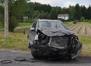 ciemne auto z rozbitym przodem stoi na poboczu drogi. w tle widać dom