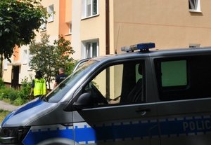 blok mieszkalny. przed blokiem stoi oznakowany radiowóz. w stronę bloku idzie dwoje umundurowanych policjantów