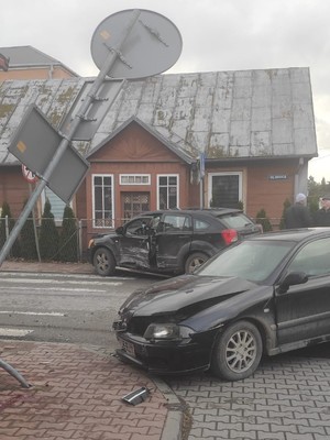 na poboczu stoi czarny samochód z rozbitym przodem. przed nim stoi wygięty znak drogowy. po drugiej stronie drogi na poboczu stoi czarny samochód z wgniecionym boikiem