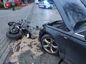 na jezdni stoi szary samochód z rozbitym przodem. przed nim leży rozbity motocykl. na jezdni widać rozlany płyn