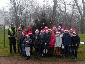 dzieci w czerwonych czapkach z białymi obwódkami i pomponami stoją przed choinką. obok dzieci stoi umundurowany policjant