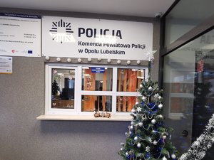 okienko dyżurki komendy Policji w Opolu lubelskim. przed okienkiem widać ustrojoną bombkami i łańcuchami choinkę. na parapecjie stoi pociąg z tworzywa imitującego piernik