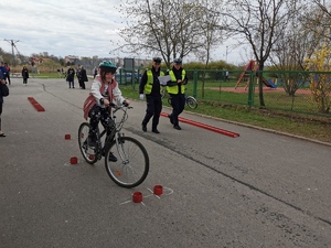 dziewczynka jhedzie na rowerze po torze przeszkód. obok niej idą policjanci z kartkami w dłoni