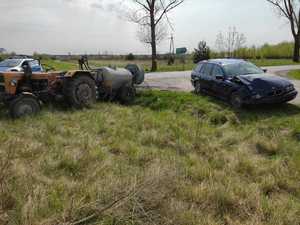 na poboczu drogi stoi ciągnik roliczy z podpiętym opryskiwaczem oraz obok niego niebieski samochód BMW z rozbitym przodem