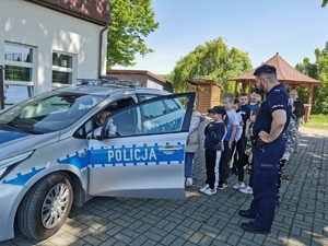 w oznakowanym radiowozie siedzi za kierownicą dziewczynka, drzwi radiowozu są uchylone. obok stoi policjant oraz grupa dzieci