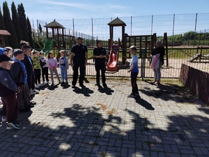 policjanci stoją na placu przed szkołą w otoczeniu dzieci. obok nich chłopiec w niebieskiej bluzce objaśnia zasady przechodzenia przez przejście dla pieszych.