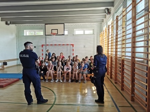 dwoje policjantów stoi przodem do siedzącej w ich stronę na ławeczkach młodzieży