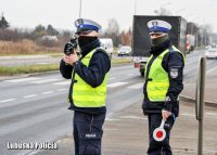 Policjant dokonuje pomiaru prędkości za pomocą ręcznego miernika. Obok stoi policjantka trzymając w dłoni tarczę sygnalizacyjną.