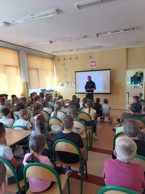 policjant w sali stoi przed siedzącą na krzesełkach młodzieżą