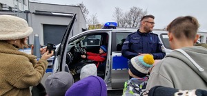 w radiowozie służbowym siedzi chłopiec w czerwonej kurtce. obok samochodu stoi policjant wraz z grupką dzieci