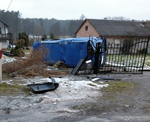 za bramą na posesji leży przewrócony niebieski bus. przed bramą widać elementy karoserii.