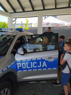 pod dachem obok radiowozu policyjnego stoi gromadka dzieci wraz z umundurowanym policjantem. część dzieci siedzi w samochodzie